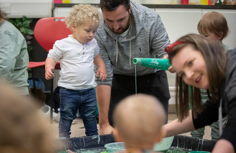 A toddler with blonde curly hair stands in a black messy tray while an adult male pours green paint into the tray in front of him. Other adults and young children are shown enjoying the mess. 
