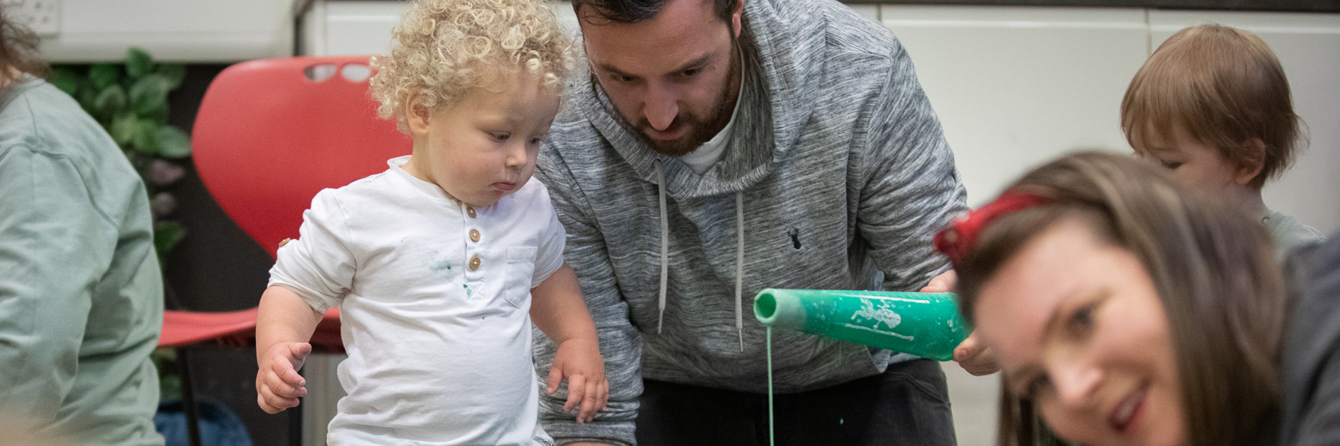 A toddler with blonde curly hair stands in a black messy tray while an adult male pours green paint into the tray in front of him. Other adults and young children are shown enjoying the mess. 