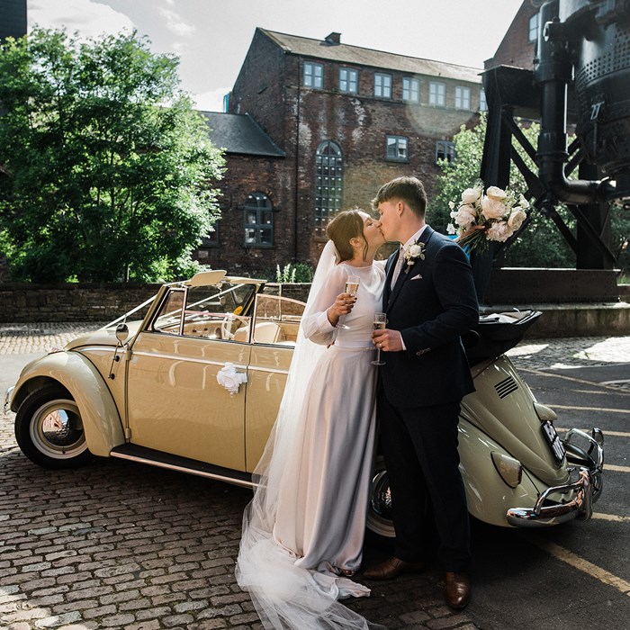 A bride in a wedding dress and a groom in a suit kiss in front a vintage car. The car is parked on a cobbled road in front of historic industrial building.