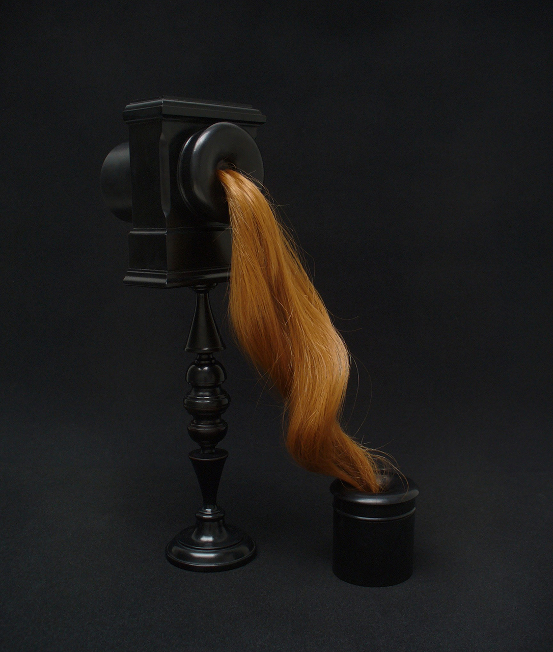 A sculpture made from repurposed ebony and human hair. A black, shiny object that looks like an old fashioned candlestick telephone receiver with long, shiny, styled, auburn hair connecting the listener and receiver.