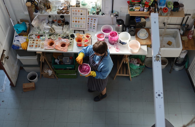 A top down photograph of an artist in their studio. Pots of dye are visible on a table and the artist is seen mixing dye.