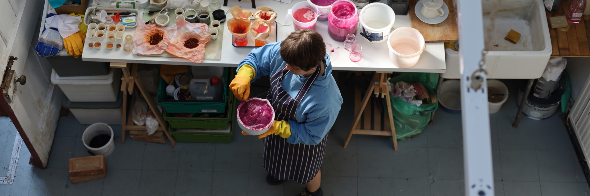 A top down photograph of an artist in their studio. Pots of dye are visible on a table and the artist is seen mixing dye.