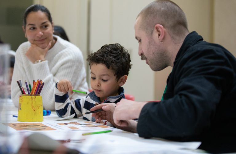 A photograph of a young boy sitting at a desk with a piece of paper in front of him and a green pencil in his hand. To the right, a woman watches on as a man instructs the child in an activity. A yellow pot of colourful pencils sits on the desk in front of them.