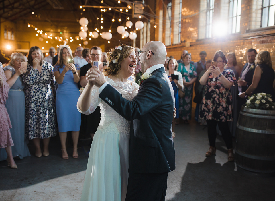 A smiling bride in a wedding dress and a groom in a suit dance in front of wedding guests in a large room decorated with fairly lights and lanterns.