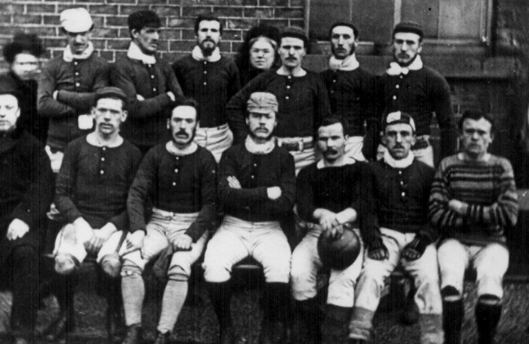 A historic black and white photograph of a group of adults. Some of the adults are wearing Victorian sporting outfits and some are wearing formal coats and hats. 