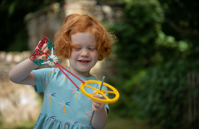 A smiling child plays with plastic cogs and wheels outside, with green foliage and tone buildings in the background
