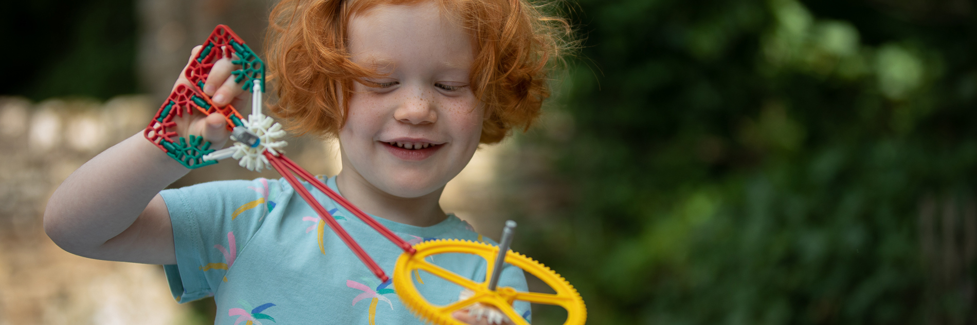 A smiling child plays with plastic cogs and wheels outside, with green foliage and tone buildings in the background