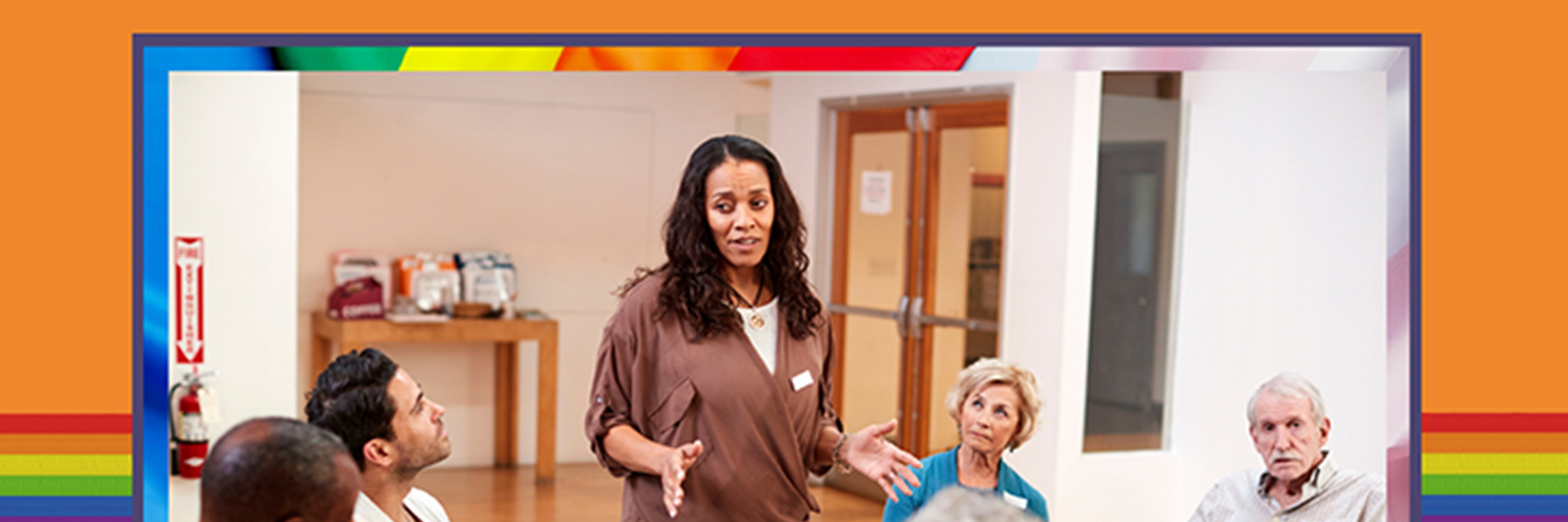 Photograph of a group of adults gathered in a circle. Five of them are seated and one is standing and talking to the group.