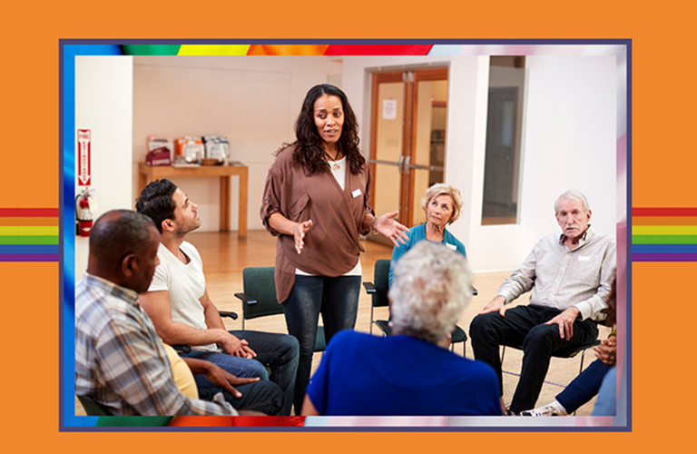 Photograph of a group of adults gathered in a circle. Five of them are seated and one is standing and talking to the group.