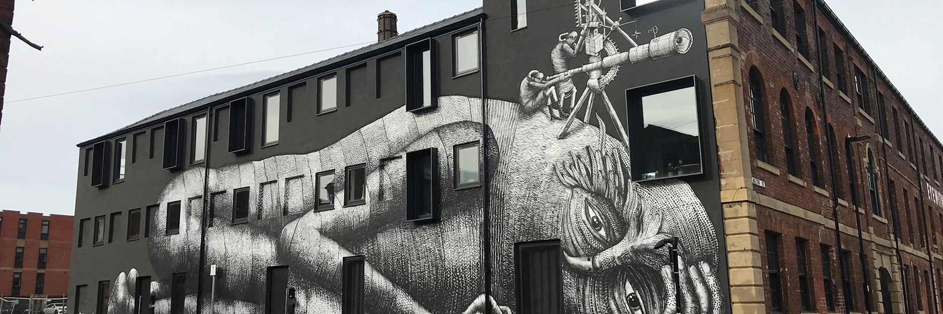 A photograph of a street art work on the side of a large building. The artwork is of a lying down giant with two human-like figures sitting on their head looking through a telescope. 