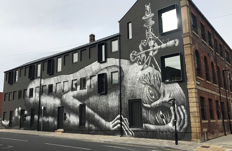A photograph of a street art work on the side of a large building. The artwork is of a lying down giant with two human-like figures sitting on their head looking through a telescope. 