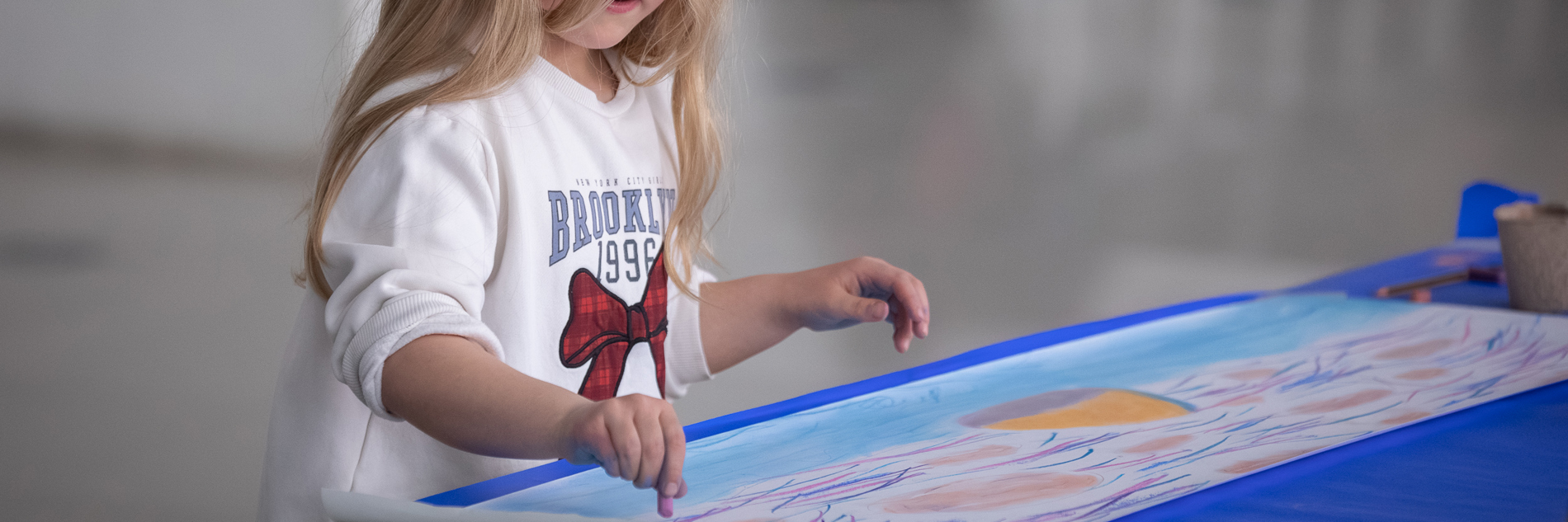 A photograph showing a young girl with long blonde hair painting a sky scene on a long piece of paper. She is wearing a white jumper with a red bow in the centre and is focused on her drawing