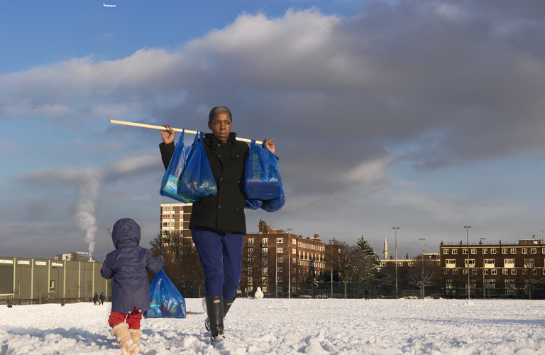 A woman with a small child beside her carries shopping bags through the snow using a poll across her shoulders