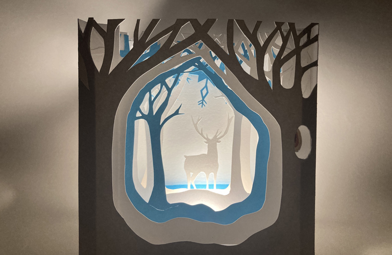 A blue, grey and black papercut scene of a stag in a forest.  