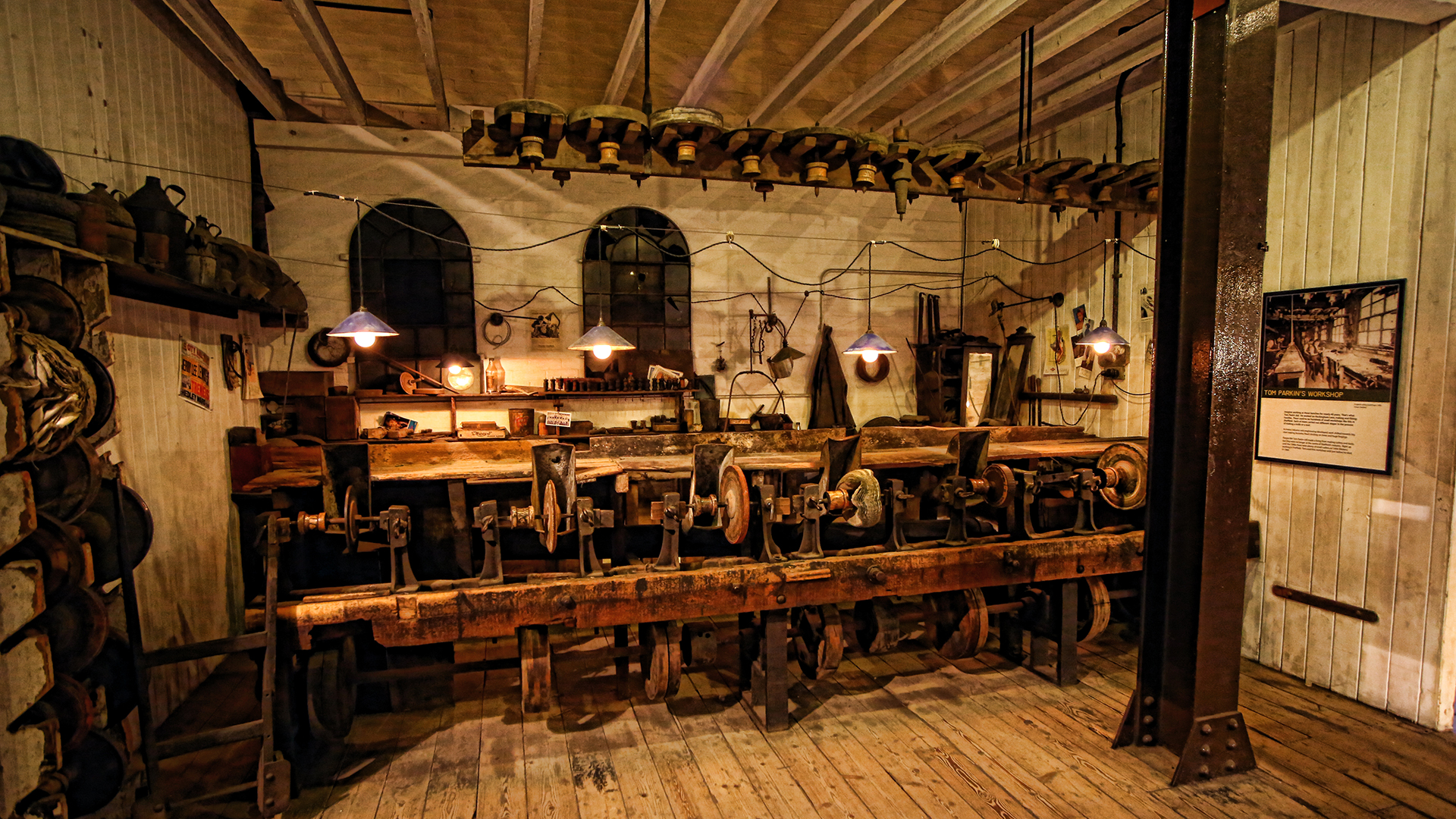 A high and well worn red brick wall fills most of the image. At ground level there are several furnace openings, now bricked up. Old tongs lean against the wall and there are several display boards including a large photograph of the workshop in its working heyday.