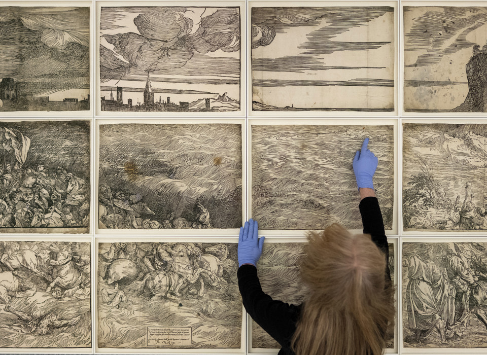 A photograph. 16 pieces of woodcut prints arranged in a grid, showing an army of men and horses being submerged in water. The back of a woman's head is seen leaning into the bottom of the frame, arms (with hands in blue gloves) stretching out over the work.