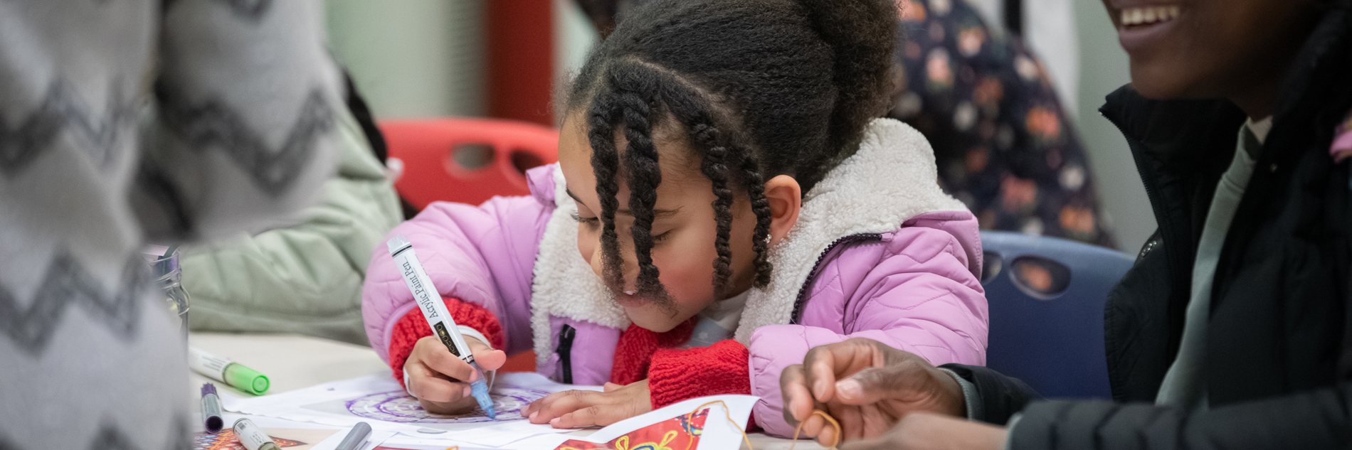 A photograph of a young girl drawing while a woman smiles in conversation with someone next to her. The little girl is focused on the colourful drawing in front of her. There are more pens on the table waiting to be used.