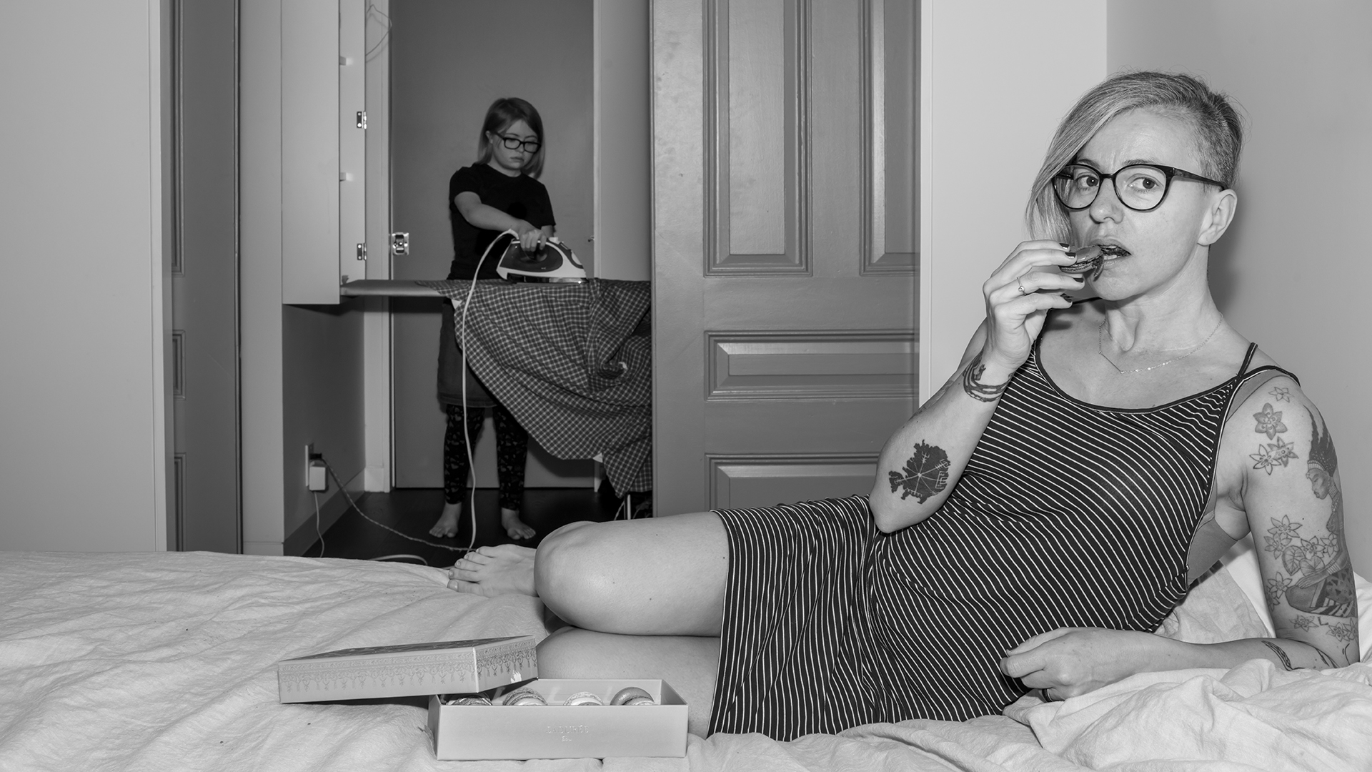 A black and white photograph of a woman eating macarons on a bed, whilst a teenage girl irons clothes in the background. 