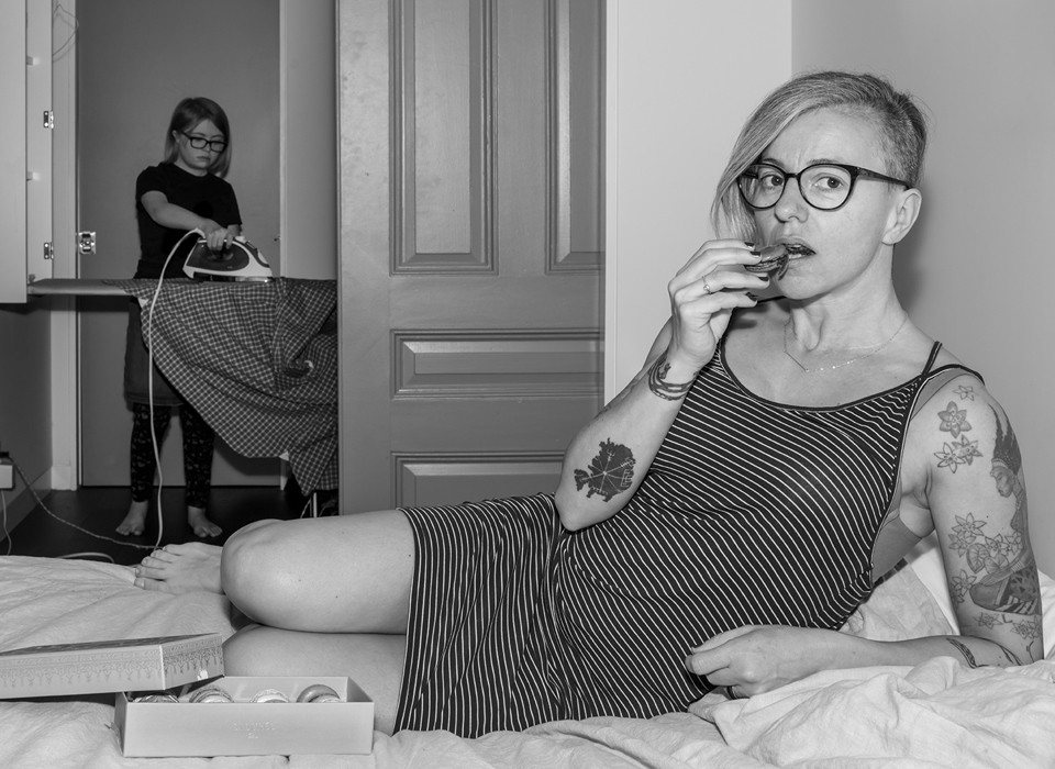 A black and white photograph of a woman eating macarons on a bed, whilst a teenage girl irons clothes in the background. 