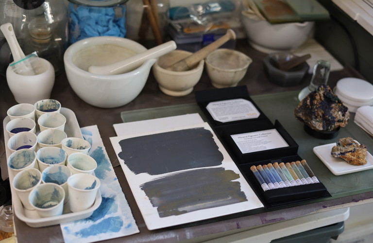 A topdown photograph of a table covered in paint samples. There are various pestle and mortars lined up on the table, and lots of paper cups with different samples of blue paint in them. Vials of ground up rocks are shown, as well as full larger rock specimens. 
