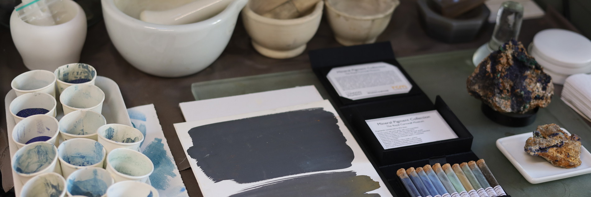 A topdown photograph of a table covered in paint samples. There are various pestle and mortars lined up on the table, and lots of paper cups with different samples of blue paint in them. Vials of ground up rocks are shown, as well as full larger rock specimens. 