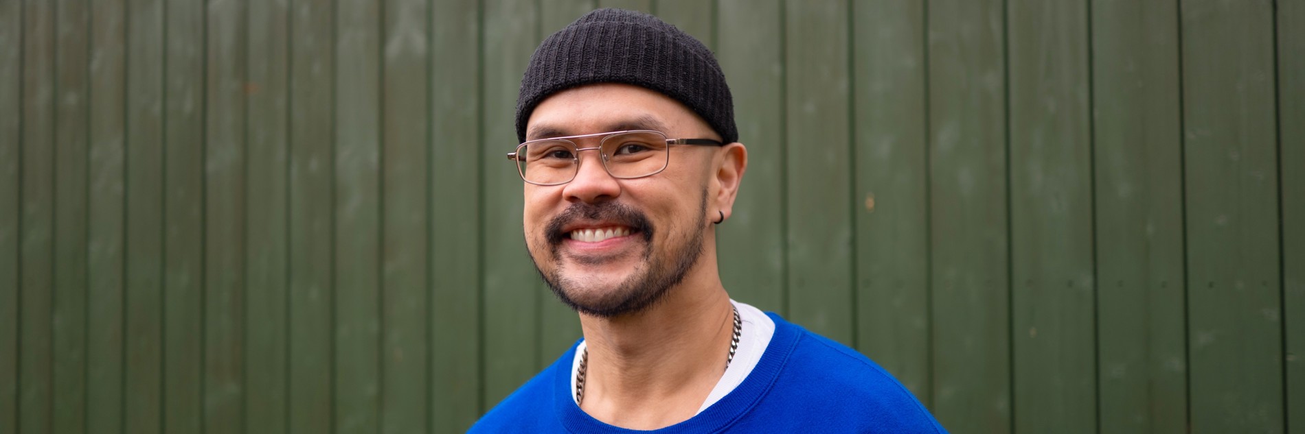 A photograph of a man in a blue jumper, black beanie hat and wire-framed glasses smiling widely at the camera. 