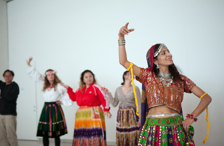 A photograph of five people dancing, with their right arms raised in the air, and their left placed on their hips. One person, probably the instructor, is shown at the front right of the image, while the others are slightly unfocused in the background. Four wear colourful skirts made out of a variety of fabrics.