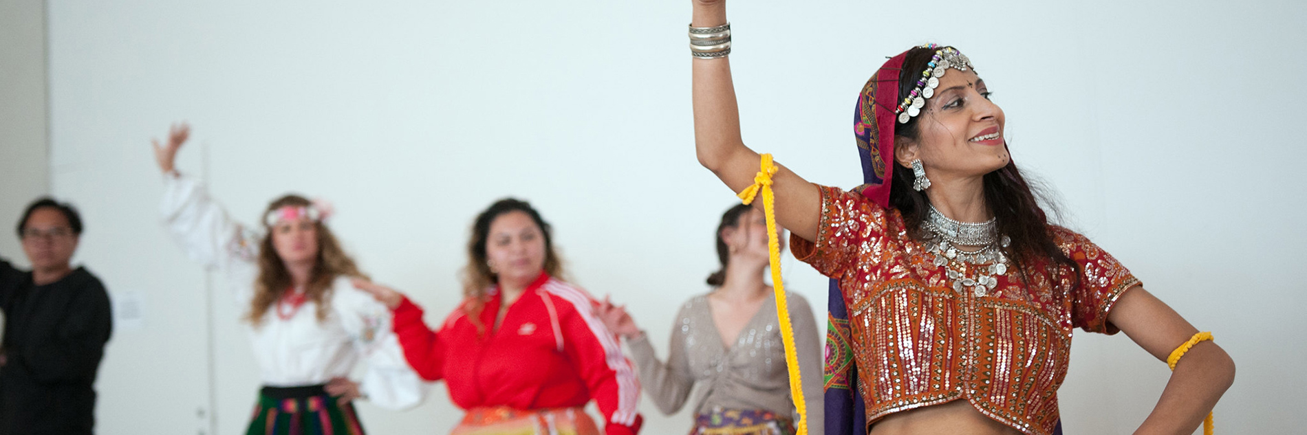 A photograph of five people dancing, with their right arms raised in the air, and their left placed on their hips. One person, probably the instructor, is shown at the front right of the image, while the others are slightly unfocused in the background. Four wear colourful skirts made out of a variety of fabrics.