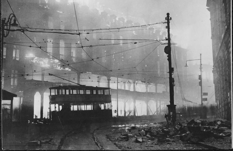 An old black and white photograph of Sheffield during the Blitz. A tram is shown off the track and there is rubble on the streets. The tram cables are visible and there is a large building behind.