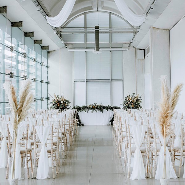 A contemporary white room with a vaulted ceiling and floor to ceiling windows on one side. Fabric covered chairs in rows face a table with floral decorations at the end of the room.