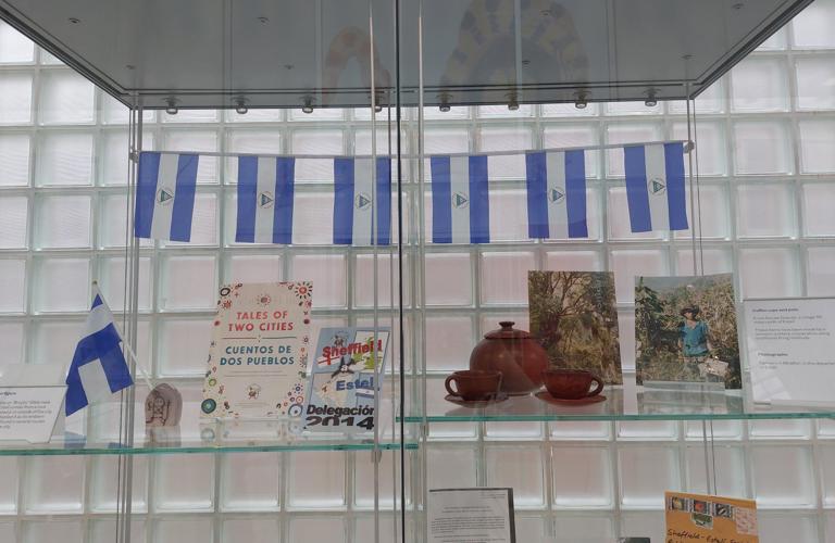 A photograph of the Sheffield Estelí display in Millennium Gallery. There are numerous objects in a glass display case. Six Nicaragua flags are shown at the top of the display. Underneath, on a shelf, are several leaflets, books and photographs. There is also a small, brown tea set, comprised of one teapot and two cups and saucers. 