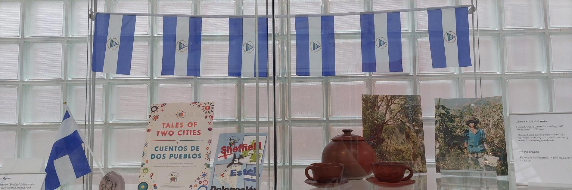 A photograph of the Sheffield Estelí display in Millennium Gallery. There are numerous objects in a glass display case. Six Nicaragua flags are shown at the top of the display. Underneath, on a shelf, are several leaflets, books and photographs. There is also a small, brown tea set, comprised of one teapot and two cups and saucers. 
