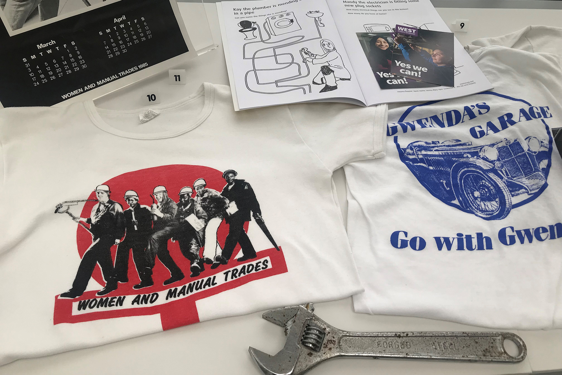 Part of a display in a museum case. There are two white t-shirts, one has a print of a blue 'Gwenda's Garage' logo. The other has a photograph of a group of women with hardhats and tools and the slogan 'Women and Manual Trades'. There is a steel spanner in front of the t-shirts.