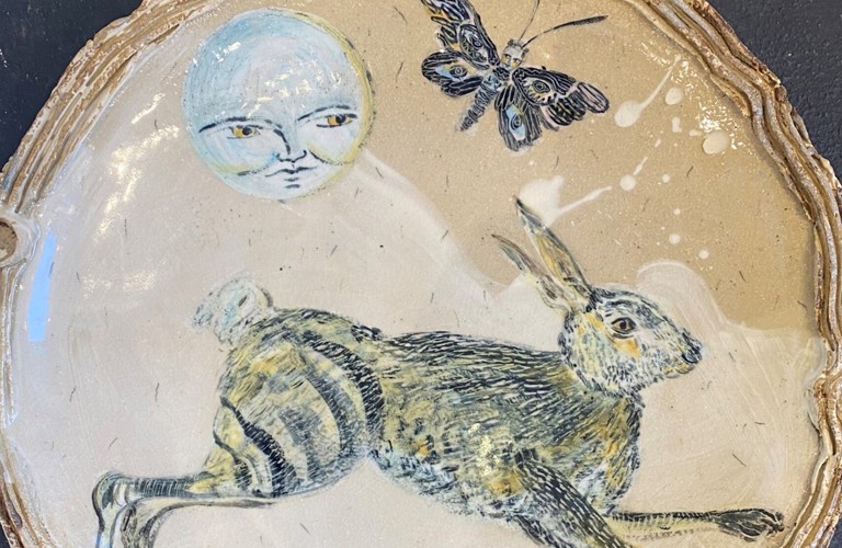 A photograph of a ceramic plate decorated with paintings of a jumping hare, a moon with a face, and a moth.