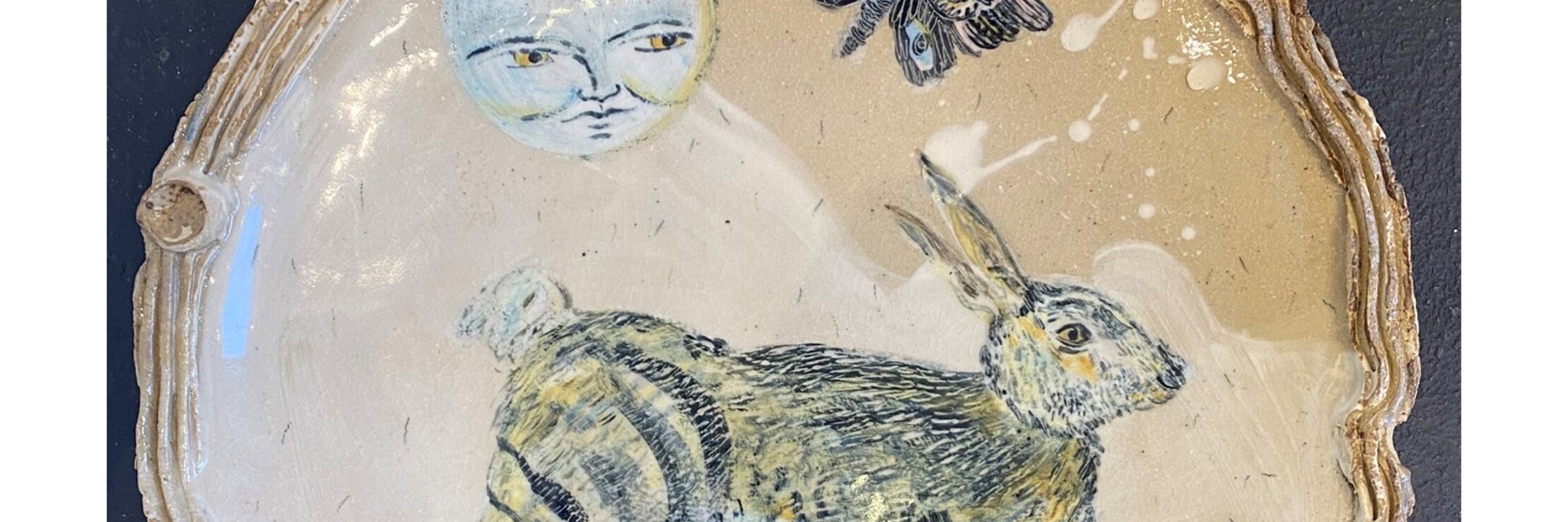 A photograph of a ceramic plate decorated with paintings of a jumping hare, a moon with a face, and a moth.