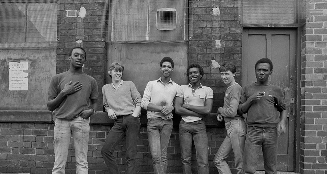 A black and white photograph of a group of young men standing in front of a red brick building.