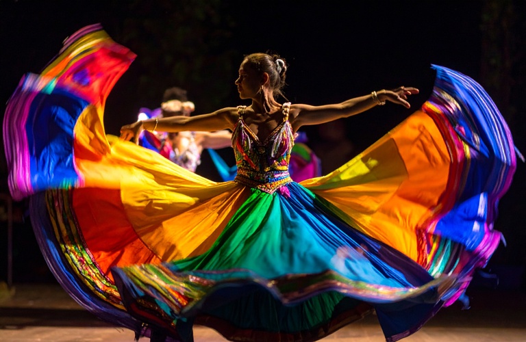 A photograph of a woman dancing against a black background, her arms outstretched. She wears a rainbow coloured skirt that flares out dramatically on all sides, filling the frame.