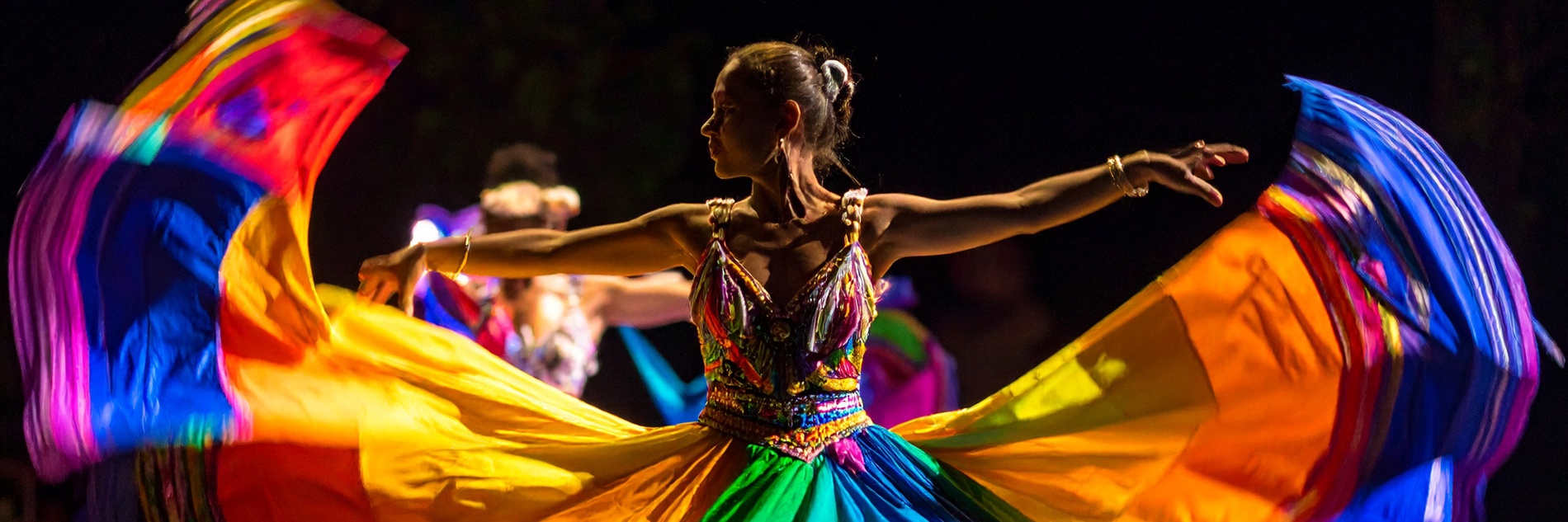 A photograph of a woman dancing against a black background, her arms outstretched. She wears a rainbow coloured skirt that flares out dramatically on all sides, filling the frame.