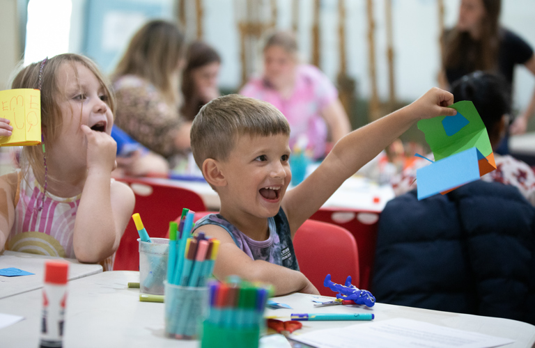 A photograph of a smiling boy holding up a piece of multicoloured, geometric artwork. Another happy child sits next to him, and there are pots of felt tips on the table in front of them