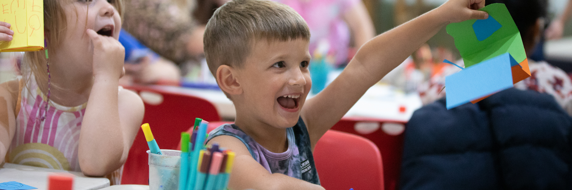 A photograph of a smiling boy holding up a piece of multicoloured, geometric artwork. Another happy child sits next to him, and there are pots of felt tips on the table in front of them