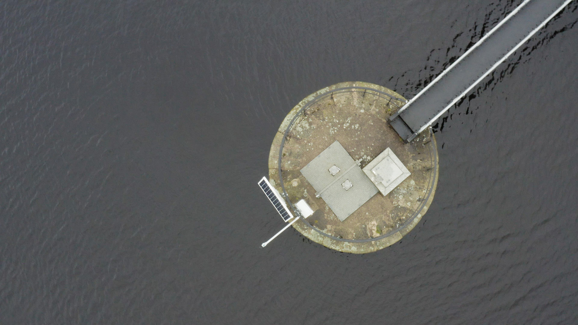 An aerial photograph of a circular concrete platform in a body of water. Extending from the platform in the top left of the image is a bridge or jetty. On the opposite side of platform to the bridge is a solar panel and in the centre is some venting and a metal panel. Around the edge of the platform is a metal railing.