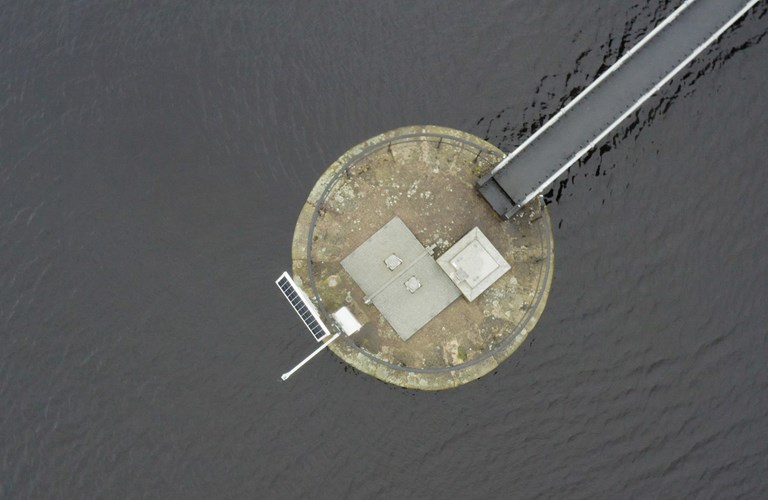 An aerial photograph of a circular concrete platform in a body of water. Extending from the platform in the top left of the image is a bridge or jetty. On the opposite side of platform to the bridge is a solar panel and in the centre is some venting and a metal panel. Around the edge of the platform is a metal railing.