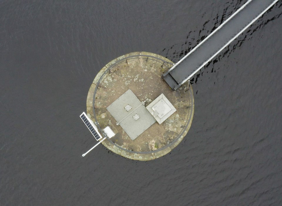 An aerial photograph of a circular concrete platform in a body of water. Extending from the platform in the top left of the image is a bridge or jetty. On the opposite side of platform to the bridge is a solar panel and in the centre is some venting and a metal panel. Around the edge of the platform is a metal railing.