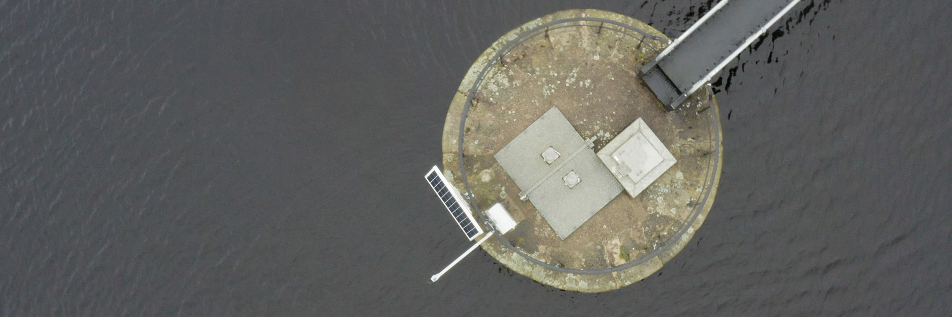 An aerial photograph of a circular concrete platform in a body of water. Extending from the platform in the top left of the image is a bridge or jetty. On the opposite side of platform to the bridge is a solar panel and in the centre is some venting and a metal panel. Around the edge of the platform is a metal railing.
