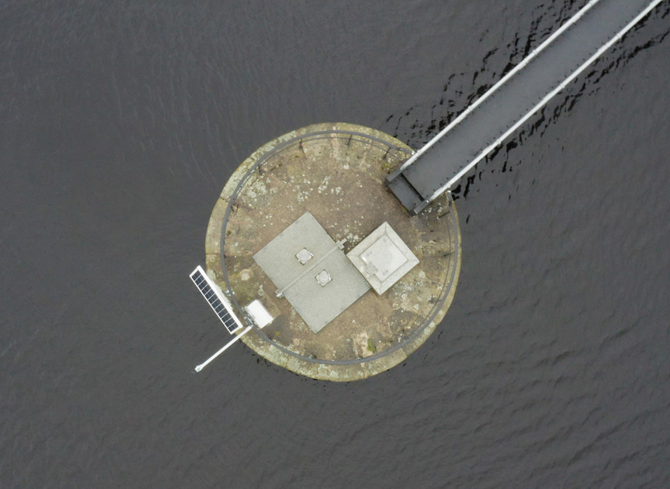 An aerial photograph of a circular concrete platform in a body of water. Extending from the platform in the top left of the image is a bridge or jetty. On the opposite side of platform to the bridge is a solar panel and in the centre is some venting and a metal panel. Around the edge of the platform is a metal railing.