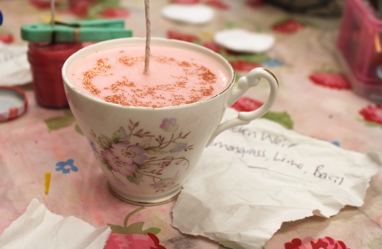 A photograph of a flowery teacup placed upon a flowery tablecloth. There is a pink candle set inside of it, with gold glitter on the surface and a fresh wick protruding from the centre. A handwritten papernote reveals the scent; lemongrass, lime and basil
