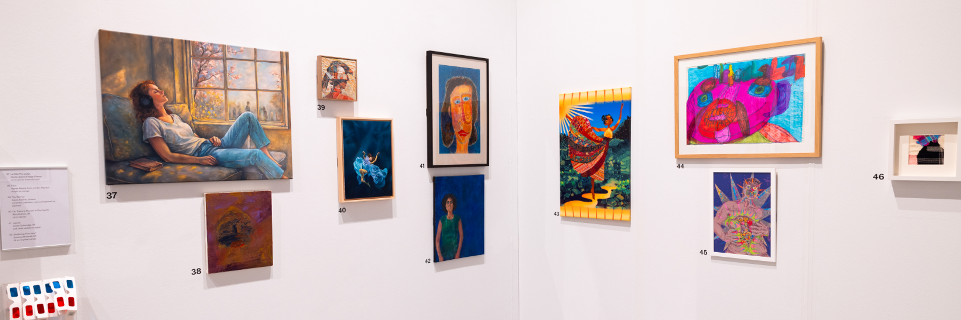 A photograph showing the corner of an exhibition. There are ten different portraits on the walls. A large one on the left of the image shows a woman relaxing in a sunlit window. Another shows a woman in a blue dress dramatically submerged in water. There is a rack of 3D glasses on the left of the wall, and barriers visible at the bottom of the walls.