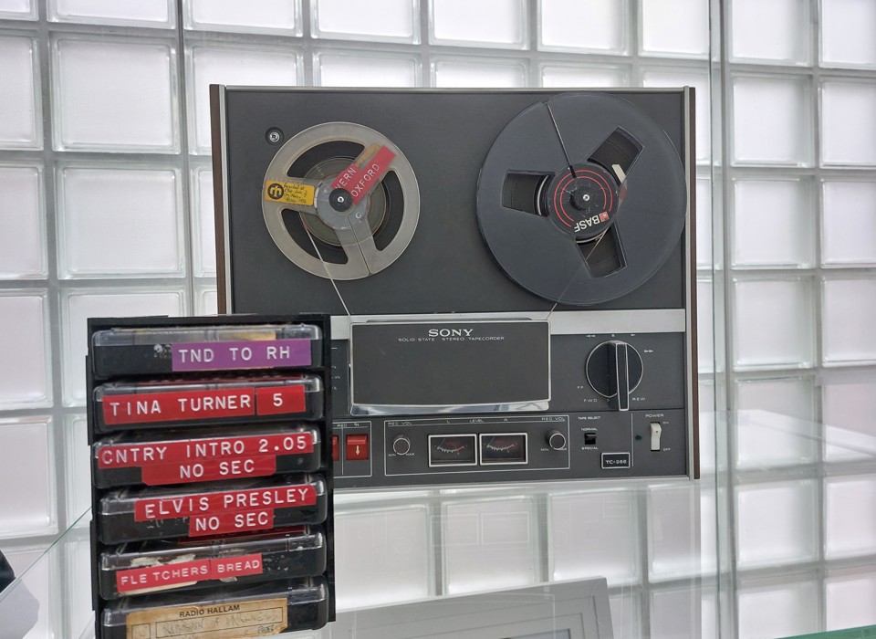A photograph of radio equipment on display in Millennium Gallery. There is a large machine with two reels in, then a stack of cassette tapes. They are labelled from top to bottom, as follows: TND TO RH, TINA TURNER 5, CNTRY INTRO 2.05 NO SEC, ELVIS PRESLEY NO SEC, and FLETCHERS BREAD