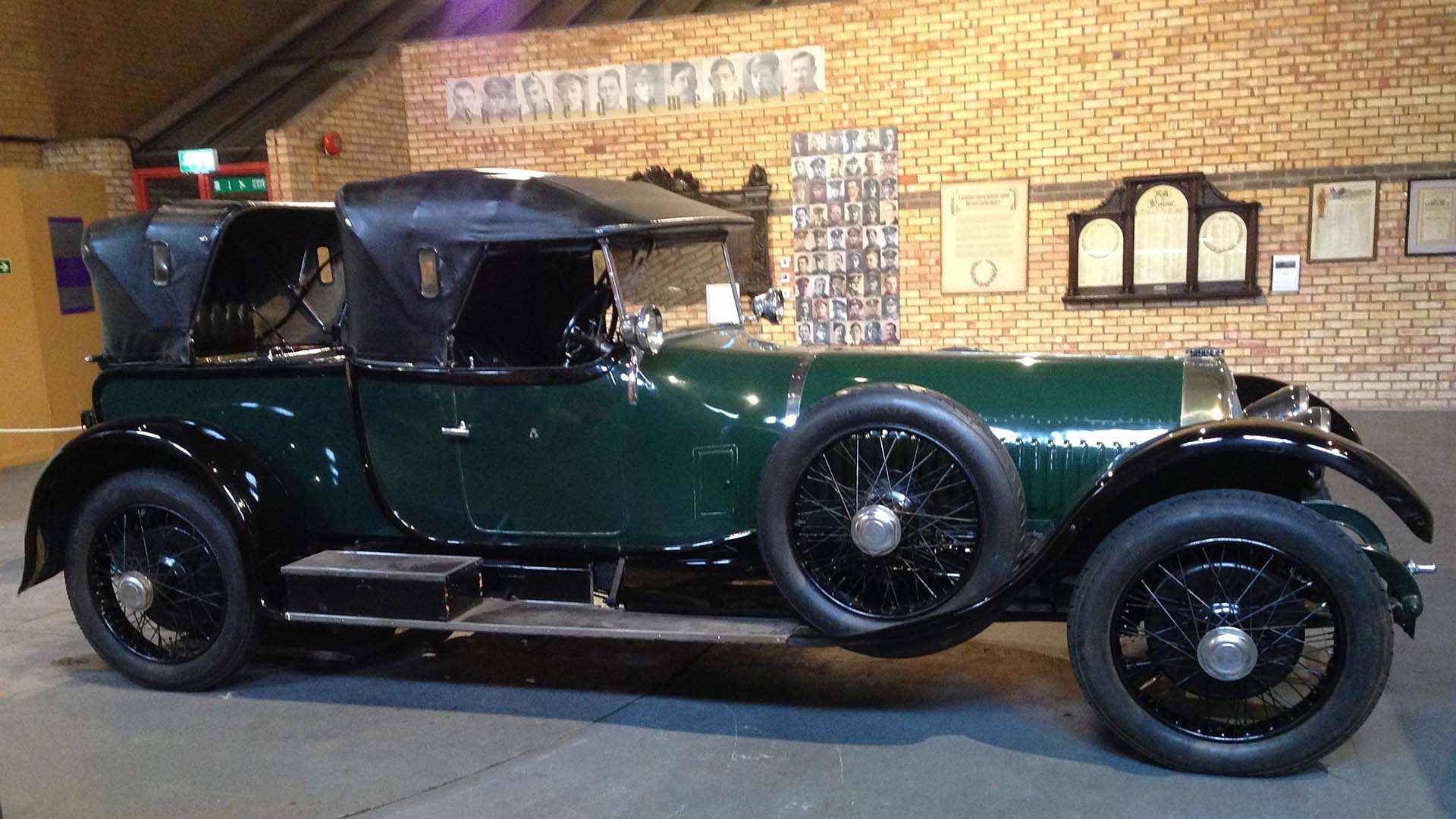 Side elevation of a large vintage car in dark green with a black leather convertible roof, chromed lamps and spoked wheels.