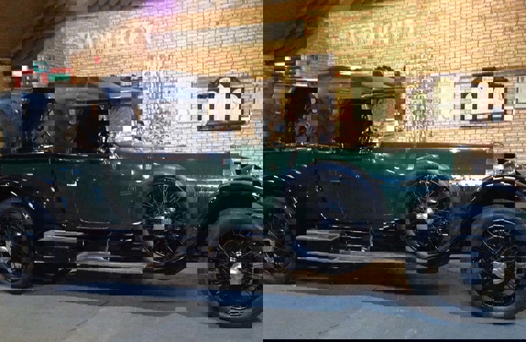 Side elevation of a large vintage car in dark green with a black leather convertible roof, chromed lamps and spoked wheels.